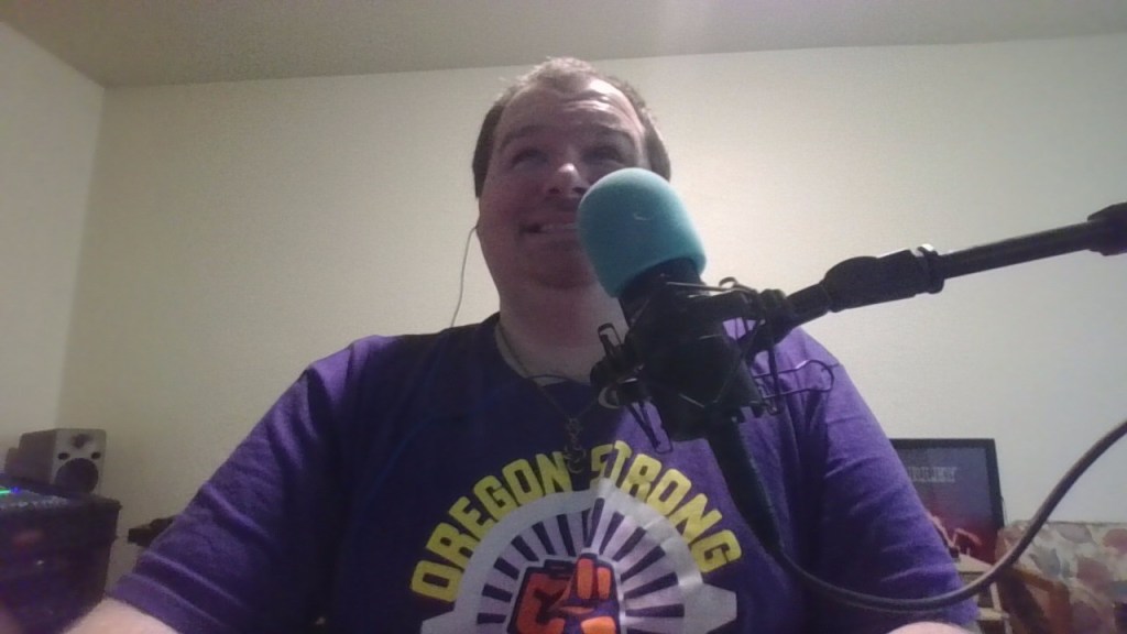 Tony is sitting in front of a microphone with a blue foam cover. He is wearing a purple shirt with the words "OREGON STRONG" in yellow letters and a graphic of a fist in the center. Tony has short hair and is smiling slightly. In the background, there is a speaker on a shelf and a framed picture or poster on the wall. The room has plain walls and a ceiling with a light source casting a soft glow.